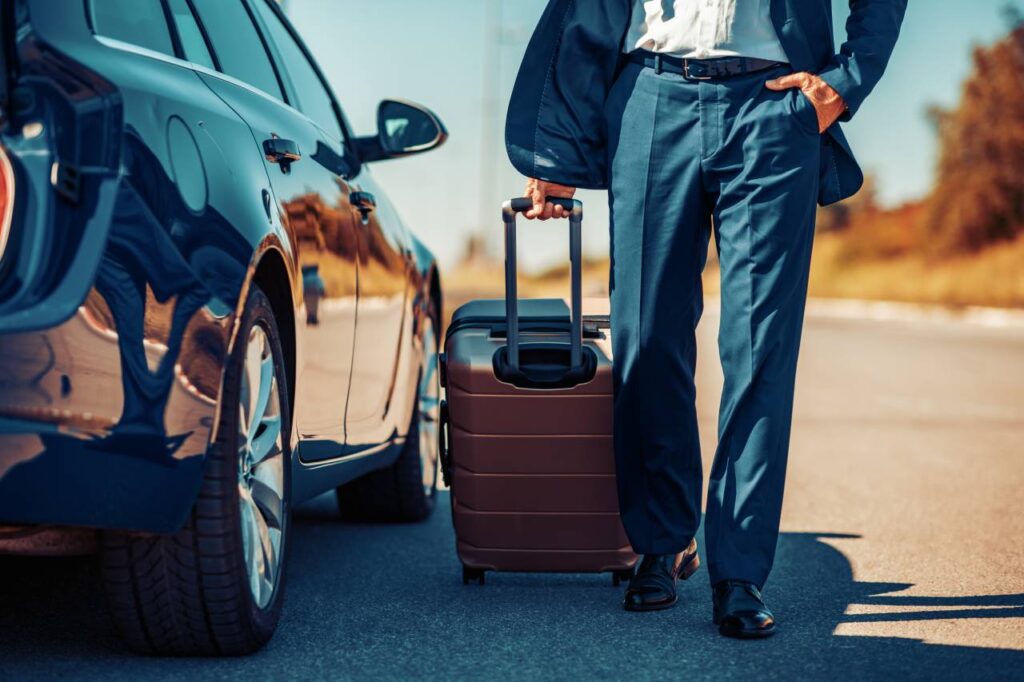 Vendeu o carro e vai viajar - bagageiro de teto carro homem carregando mala de rodinhas perto de um carro