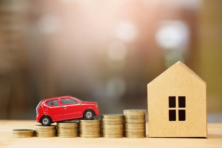 Manual do Comprador 5 Miniatura de carro vermelho sobre pilhas de moedas ao lado de uma casa de papelão, simbolizando financiamento; ideal para ilustrar conteúdos sobre o melhor banco para financiar veículo