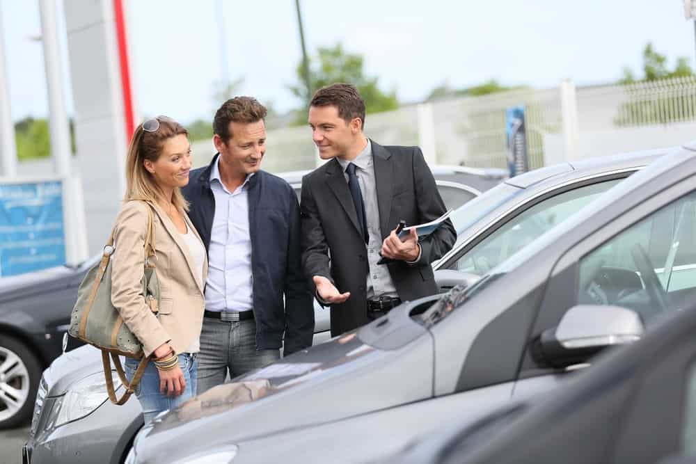 Uma mulher e dois homens vendo um carro. Um desses homens é o vendedor mostrando o carro e a mulher e o outro homem os compradores.