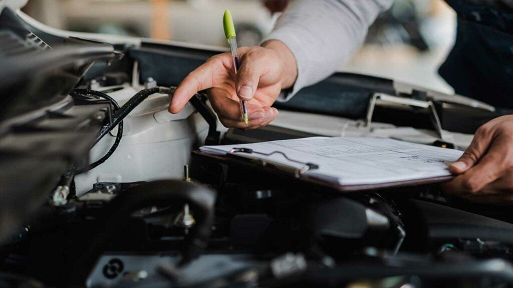Mãos segurando uma prancheta e uma caneta analisando e fazendo anotações sobre o motor de um veículo - vender carro pessoa física