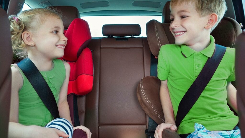 Duas crianças no banco de trás de um veículo. Ambas usando o cinto de segurança. 